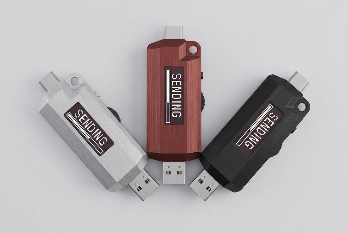 Three Lomenett offline password typers arranged in a fan: natural aluminum, dried-blood red, and matte black anodized finishes, each screen showing SENDING