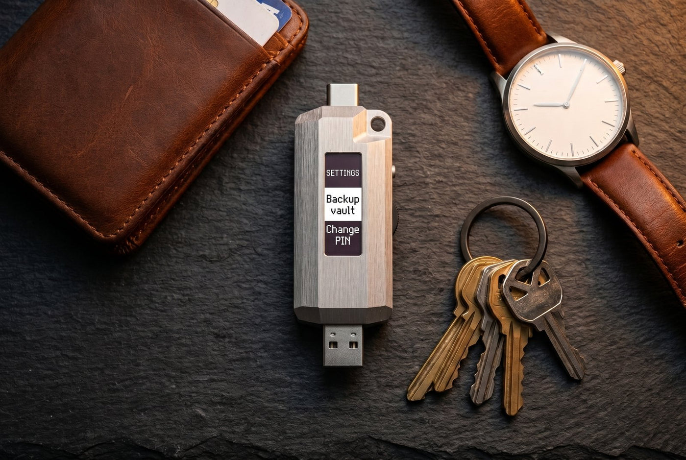 Lomenett laid out with a wallet, keys and a watch on slate, screen showing the settings menu; everyday carry for an offline password manager