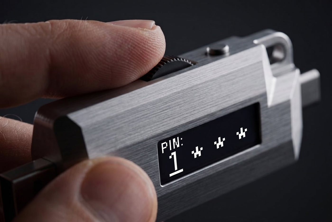 Macro photo of a thumb on the Lomenett thumbwheel while the 128 by 32 OLED shows PIN entry; the PIN is entered on the offline hardware password manager itself, never on the computer